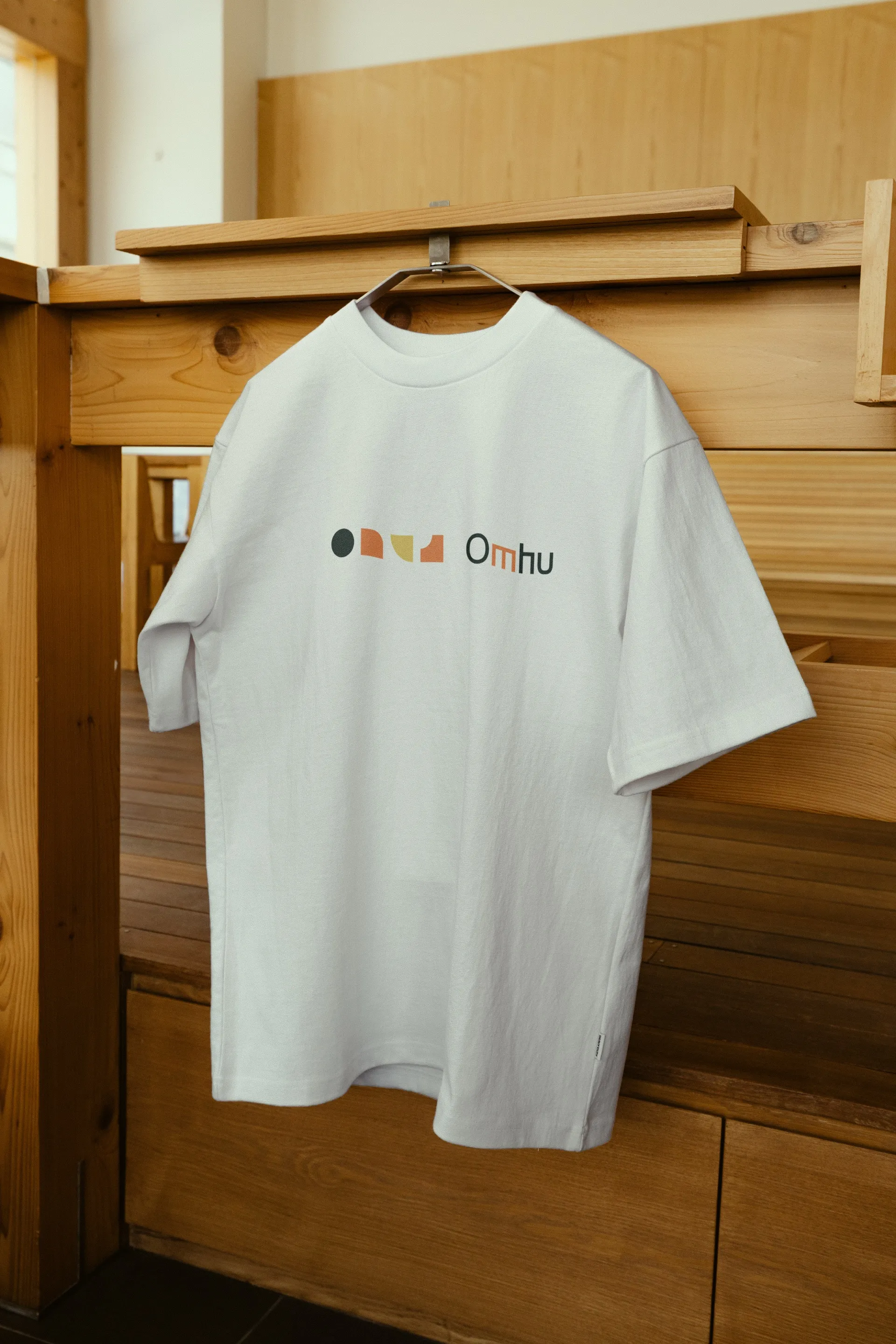 A white T-shirt with Omhu’s shapes and logo, hanging in a wooden room